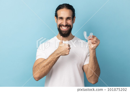 Portrait of man with beard wearing white T-shirt holding dental aligner retainer, dental clinic for beautiful teeth, showing thumb up. Indoor studio shot isolated on blue background. Portrait of man with beard wearing white T-shirt holding dental aligner retainer, dental clinic for beautiful teeth, showing thumb up. Indoor studio shot isolated on blue background. 101875202