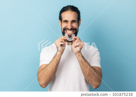 Portrait of man with beard in white T-shirt using teeth whitening braces, dental aligner retainer, dental clinic for beautiful teeth, treatment course. Indoor studio shot isolated on blue background. 101875238