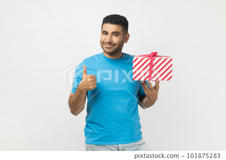 Portrait of attractive joyful unshaven man wearing blue T- shirt standing holding wrapped present box, showing like gesture, thumb up, congratulating. Indoor studio shot isolated on gray background. 101875283