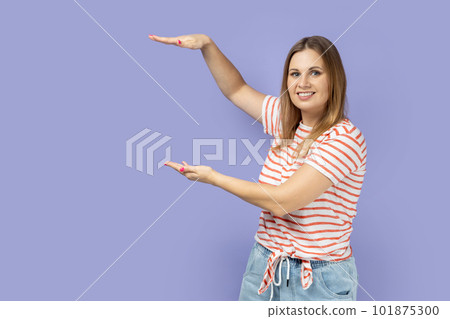 Portrait of satisfied smiling blond woman wearing striped T-shirt presenting area between hands for advertisement, showing huge size. Indoor studio shot isolated on purple background. Portrait of satisfied smiling blond woman wearing striped T-shirt presenting area between hands for advertisement, showing huge size. Indoor studio shot isolated on purple background. 101875300