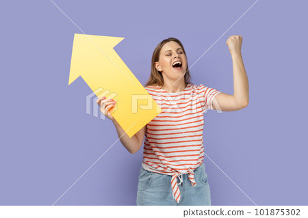 Portrait of extremely happy woman wearing striped T-shirt holding big yellow arrow pointing aside and clenched fist, celebrating victory, increase. Indoor studio shot isolated on purple background. Portrait of extremely happy woman wearing striped T-shirt holding big yellow arrow pointing aside and clenched fist, celebrating victory, increase. Indoor studio shot isolated on purple background. 101875302