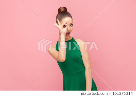Portrait of funny childish young adult woman standing covering her eye with ok gesture, looking at camera, showing okay sign, wearing green dress. Indoor studio shot isolated on pink background. Portrait of funny childish young adult woman standing covering her eye with ok gesture, looking at camera, showing okay sign, wearing green dress. Indoor studio shot isolated on pink background. 101875314