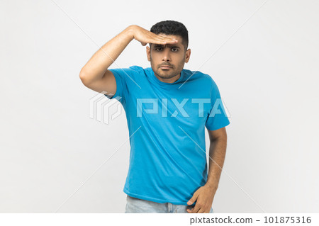 Portrait of unshaven man wearing blue T- shirt standing keeps hand near forehead, looks far away, searches something on horizon. Indoor studio shot isolated on gray background. 101875316