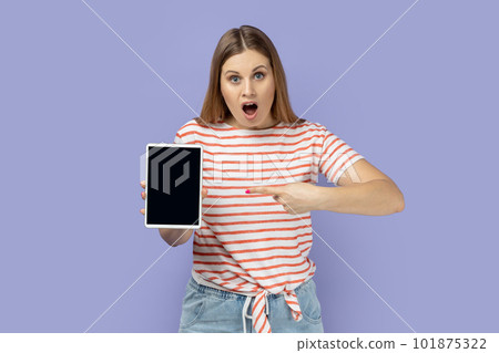 Portrait of amazed shocked surprised blond woman holding pointing at tablet with empty display, mockup screen for your advertisement. Indoor studio shot isolated on purple background. 101875322