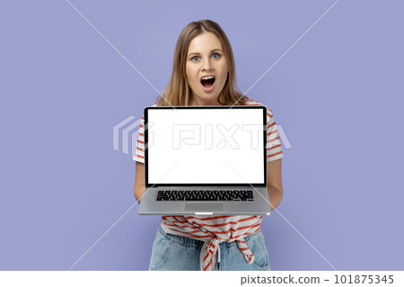 Portrait of shocked amazed blond woman wearing striped T-shirt holding laptop with white empty display with empty space for advertisement. Indoor studio shot isolated on purple background. 101875345