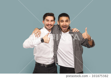 Portrait of two satisfied men friends, standing together and hugging, showing thumbs up, recommend service, good feedback, Indoor studio shot isolated on light blue background. Portrait of two satisfied men friends, standing together and hugging, showing thumbs up, recommend service, good feedback, Indoor studio shot isolated on light blue background. 101875382