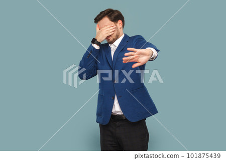 Portrait of man with mustache standing and covering eyes with palm, showing stop gesture, doesn't want to see, wearing official style suit. Indoor studio shot isolated on light blue background. Portrait of man with mustache standing and covering eyes with palm, showing stop gesture, doesn't want to see, wearing official style suit. Indoor studio shot isolated on light blue background. 101875439