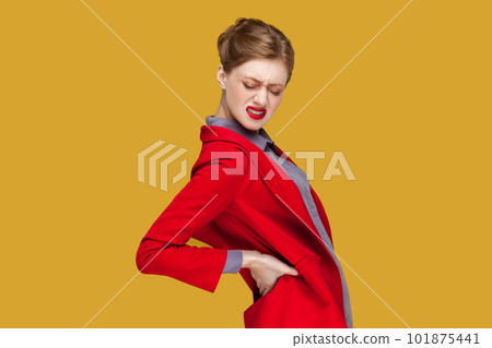 Side view portrait of sad sick unhealthy woman with red lips standing with frowning face, touching her painful back, wearing red jacket. Indoor studio shot isolated on yellow background. 101875441