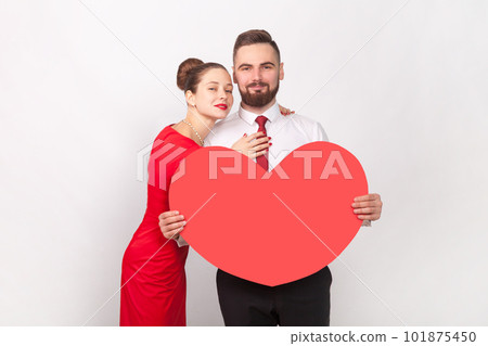 Portrait of pretty pleased loving couple in elegant attires standing together and hugging, man holding big heart, looking at camera. Indoor studio shot isolated on gray background. 101875450