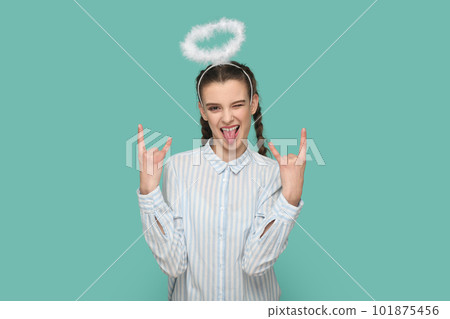 Portrait of extremely happy teenager girl with braids wearing striped shirt and nimb over her hair, showing rock and roll gesture, showing tongue. Indoor studio shot isolated on green background. 101875456