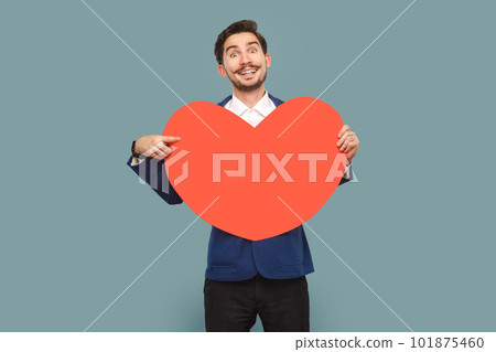 Portrait of handsome man with mustache standing holding pointing at big red heart, smiling toothily to camera, wearing white shirt and jacket. Indoor studio shot isolated on light blue background. Portrait of handsome man with mustache standing holding pointing at big red heart, smiling toothily to camera, wearing white shirt and jacket. Indoor studio shot isolated on light blue background. 101875460