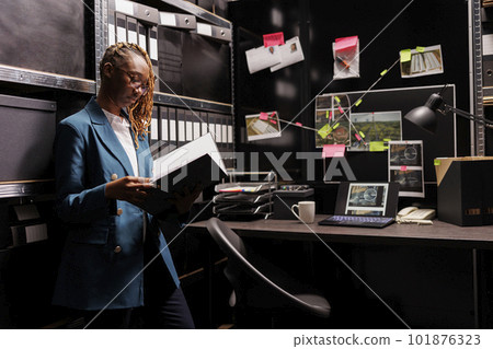 African american woman police investigator reading case file in office room. Private detective holding folder and analyzing investigation report near board with clues connection scheme African american woman police investigator reading case file in office room. Private detective holding folder and analyzing investigation report near board with clues connection scheme 101876323