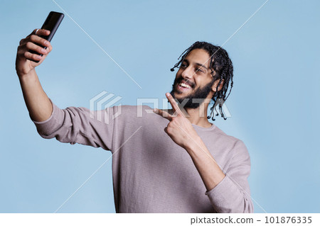 Young arab man smiling while holding mobile phone during video call and showing peace gesture with fingers. Cheerful person looking at smartphone front camera while taking selfie Young arab man smiling while holding mobile phone during video call and showing peace gesture with fingers. Cheerful person looking at smartphone front camera while taking selfie 101876335