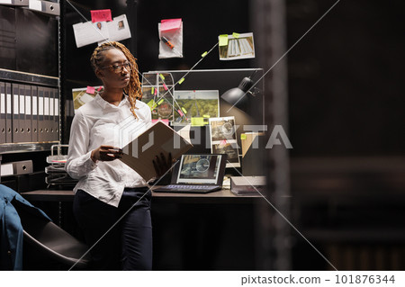 Police detective reading crime case file and thinking about clues late. Law enforcement investigator holding forensic expertise report while standing at dark office workplace desk 101876344