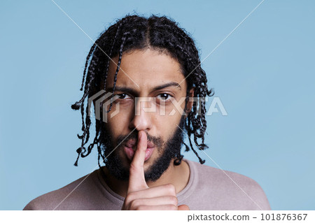 Arab man making silent gesture with serious facial expression and looking at camera. Young person with raised eyebrow showing quiet sign while holding finger on lips portrait 101876367