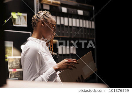 Thoughtful african american woman detective reading criminal investigation documentation in agency office room. Law enforcement professional studying criminologist report at night 101876369