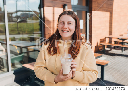 Happy girl drinking milkshake in the street. Cheerful young woman wearing yellow hoodie walking outdoors. Urban lifestyle concept. Drink to go. Enjoy moment, take a break. Happy girl drinking milkshake in the street. Cheerful young woman wearing yellow hoodie walking outdoors. Urban lifestyle concept. Drink to go. Enjoy moment, take a break. 101876370