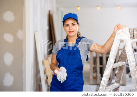 Portrait of positive builder woman in blue overalls next to stepladder 101880300