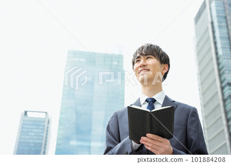 A man in his 20s and 30s in a suit looking into the distance with a notebook in his hand in a business district 101884208