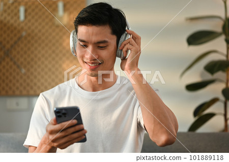 Joyful millennial asian man enjoying listening to favorite music online on headphone and using smartphone in living room Joyful millennial asian man enjoying listening to favorite music online on headphone and using smartphone in living room 101889118