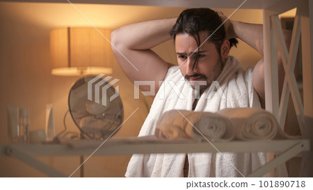 Grooming routine. Tidy man. Freshness hygiene. Handsome shirtless guy with mustache tying up long dark hair looking in mirror in bathroom interior. 101890718