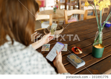 Beautiful young woman is guessing on cards with tarot, runes on wooden table and uses an online app in phone to interpret divination, home interior,candles and dry herbs.. Beautiful young woman is guessing on cards with tarot, runes on wooden table and uses an online app in phone to interpret divination, home interior,candles and dry herbs.. 101891776