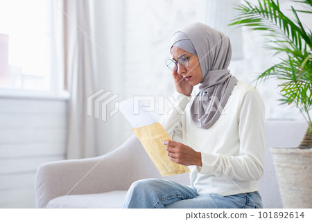 Muslim woman at home in hijab upset, arab woman received mail message notification with bad news, woman reading pensive and worried sitting on sofa in living room. 101892614