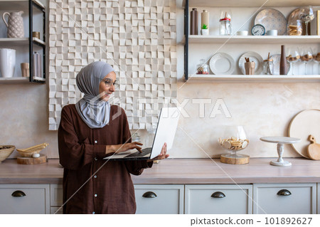 Joyful and successful business woman in hijab at home using laptop while standing, muslim woman working remotely. 101892627