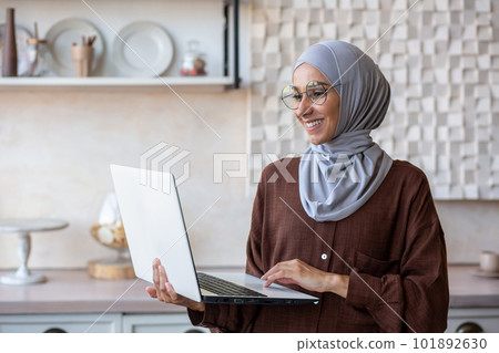 Joyful and successful business woman in hijab at home using laptop while standing, muslim woman working remotely. 101892630