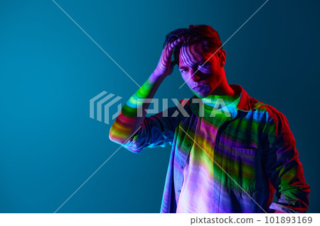 Handsome young man with multi colored neon digital stripes reflection standing with serious expression over blue background Handsome young man with multi colored neon digital stripes reflection standing with serious expression over blue background 101893169