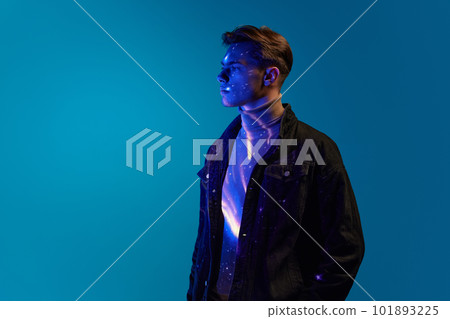 Portrait of handsome young man in stylish clothes, posing with serious expression over blue background in neon light. Digital light reflection on body Portrait of handsome young man in stylish clothes, posing with serious expression over blue background in neon light. Digital light reflection on body 101893225