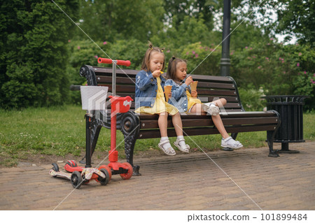 Two little sisters of preschool age sat down on a bench in the park to relax and eat a cool ice cream cone. The children are tired of riding scooters and hungry 101899484
