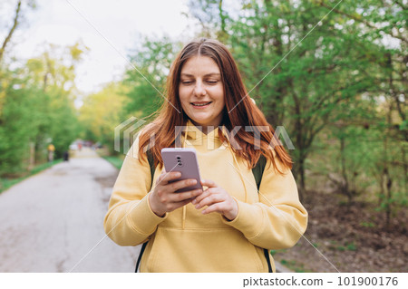 A portrait of a smiling beautiful woman walking with phone on nature background. Happy woman with backpack is using a smartphone in park outdoors, spring time. Traveler 101900176
