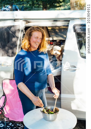 Middle aged nomad woman cooking salad outside her camper van in the forest camp with her cockapoo puppy pet during road trip. Best friend. Enjoying free lifestyle, travel vacation. Travel with pets. Middle aged nomad woman cooking salad outside her camper van in the forest camp with her cockapoo puppy pet during road trip. Best friend. Enjoying free lifestyle, travel vacation. Travel with pets. 101904911