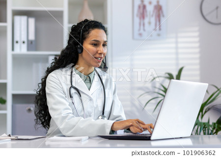 Successful hispanic female doctor working inside medical office at desk, woman typing on laptop, smiling with video call headset, female doctor consulting patients remotely online. 101906362