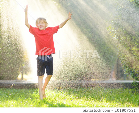 Funny little boy playing during a rain in sunny city park. Elementary school child laughing, jumping and having fun with spray of water. Summer outdoors activity for kids. Funny little boy playing during a rain in sunny city park. Elementary school child laughing, jumping and having fun with spray of water. Summer outdoors activity for kids. 101907551