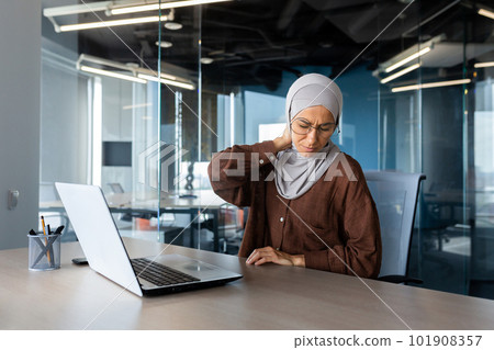 Tired young arab businesswoman in hijab working on project overtime in office at laptop and holding neck, grimacing in pain, massaging with hands. 101908357