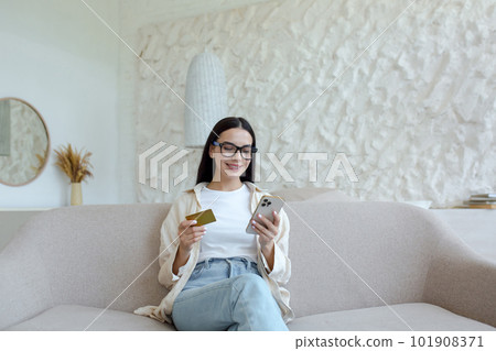 A young woman is sitting on the sofa at home and using the phone. Holds a credit card and makes remote purchases, rests, has fun. 101908371