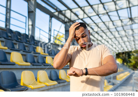 Upset hispanic sports man standing in stadium, holding head and looking at bracelet, smart watch. Dissatisfied with the training result and running time. Upset hispanic sports man standing in stadium, holding head and looking at bracelet, smart watch. Dissatisfied with the training result and running time. 101908448