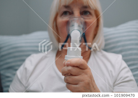 Portrait of sick senior woman sitting with an inhaler. Unhealthy female doing inhalation at home, she use nebulizer and inhaler for the treatment, sitting on the couch at home, need treatment. Portrait of sick senior woman sitting with an inhaler. Unhealthy female doing inhalation at home, she use nebulizer and inhaler for the treatment, sitting on the couch at home, need treatment. 101909470