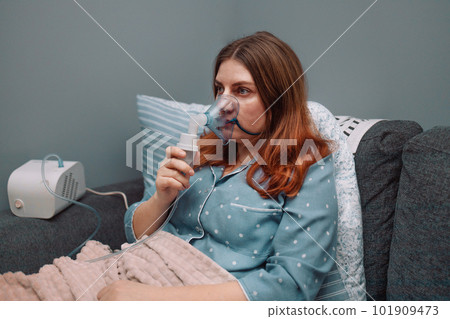 Young sick woman putting on nebulizer mask on face to make inhalation, using an inhaler at home. Medical inhaler emergency equipment, asthmatic attack 101909473