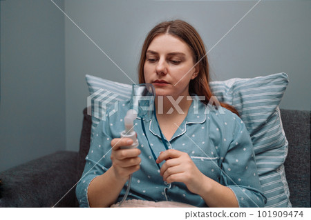 Young sick woman putting on nebulizer mask on face to make inhalation, using an inhaler at home. Medical inhaler emergency equipment, asthmatic attack 101909474