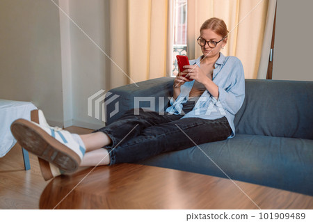 Blonde 30s girl checking social media holding smartphone at home. Smiling young blonde woman using mobile phone app playing game, shopping online, ordering delivery relax on sofa. 101909489