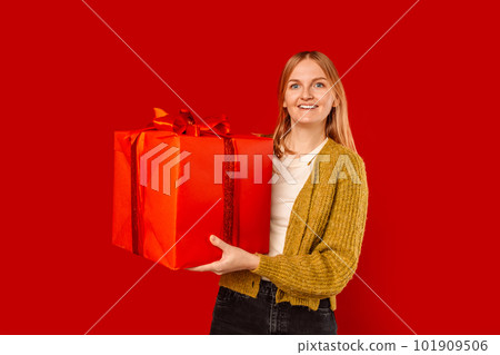 Smiling beautiful young blonde caucasian woman standing hold big red present box with gift ribbon bow looking camera isolated on bright red color background, studio portrait 101909506