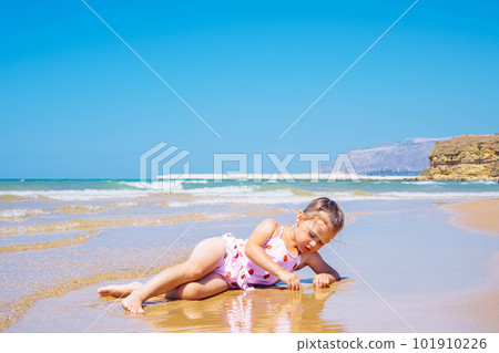 Cute little girl in pink swimsuit with strawberries lying in sand and play with water at the seashore against blue sky 101910226