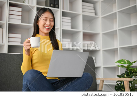Adorable young woman using laptop and drinking coffee while sitting on the sofa 101920513