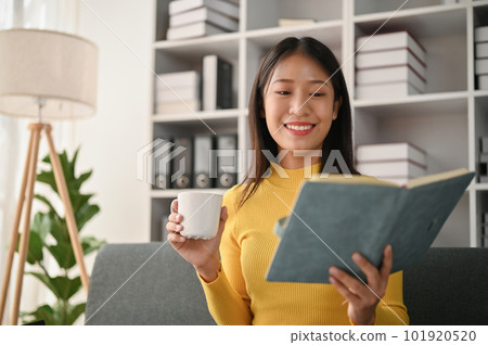 Young Asian woman reading book in her living room while drinking a coffee 101920520