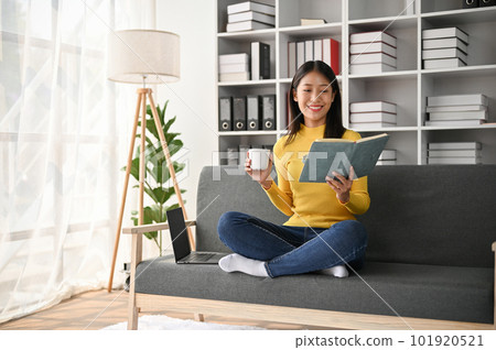 Adorable young Asian girl drinking a cup of coffee while reading book on sofa Adorable young Asian girl drinking a cup of coffee while reading book on sofa 101920521