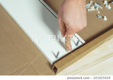 Male hands finger wooden dowel to fix walls of white drawer, assembling dresser or wardrobe. Eccentric furniture ties 101925029