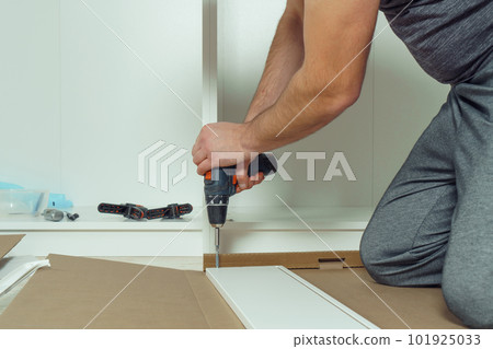 Male hands hold cordless electric screwdriver, screw to fix walls of white drawer, assembling dresser, wardrobe on floor Male hands hold cordless electric screwdriver, screw to fix walls of white drawer, assembling dresser, wardrobe on floor 101925033
